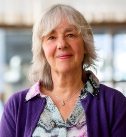 A woman with shoulder-length, light grey hair looks at the camera. She is wearing a purple cardigan over a printed blouse