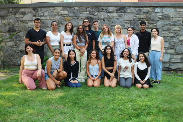 Two rows of smiling people, some standing and some kneeling, on a lawn in front of a brick wall