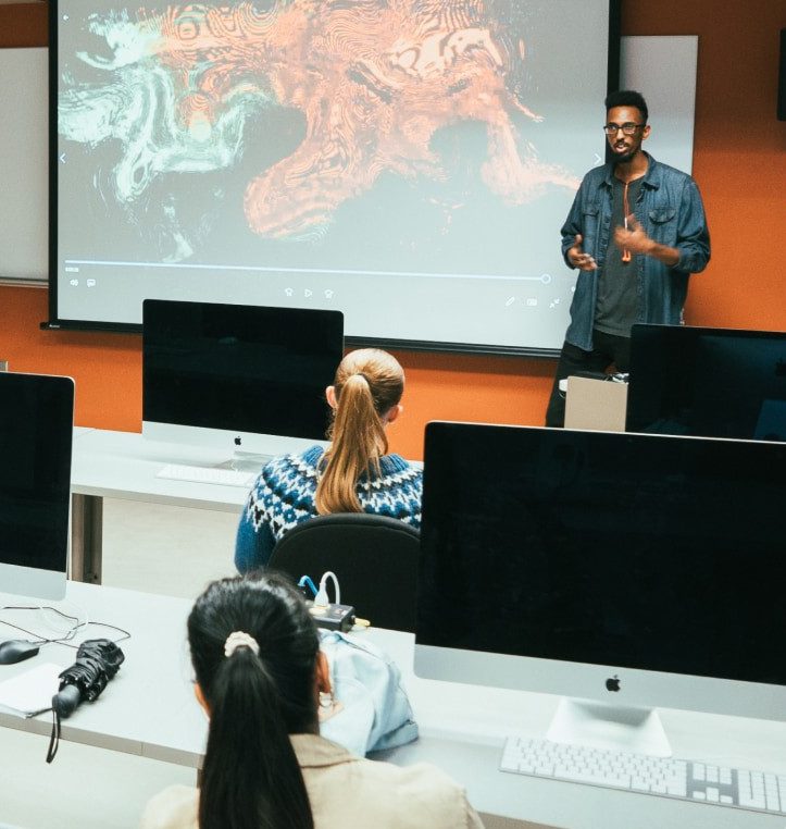 Students are sitting in a classroom with computers, listening to an in-person presentation