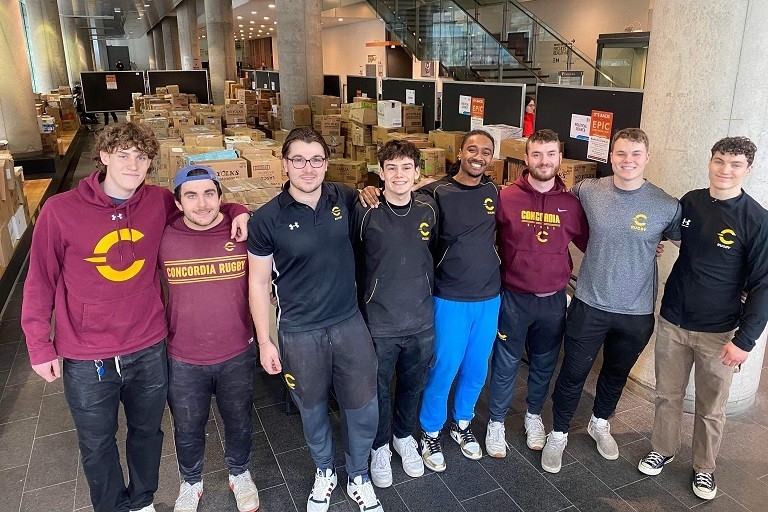 Members of the men’s rugby team stand in front of rows of cardboard boxes filled with used books.