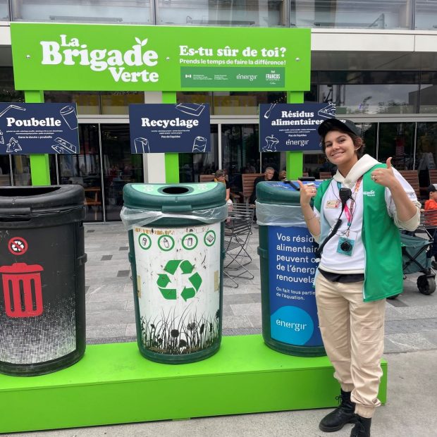 A photograph of an individual giving a thumbs up in front of recycling bins.