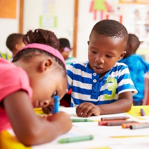 A photograph of two Black children writing with crayons in a classroom.