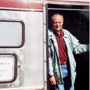  The image features Horst Hutter standing in the doorway of a train car. He is casually dressed, with a light-colored jacket over a dark red shirt, blue jeans, and a belt. His expression is relaxed and content, indicative of a moment of leisure or travel.