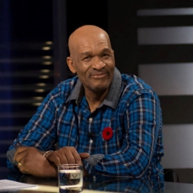  The image is a candid shot of Egbert Gaye seated and smiling. He is wearing a blue and black checked shirt and a grey vest, and there is a red poppy pin on his left lapel. He has a glass of water in front of him on the table, indicating he may be in an interview setting. The background is out of focus with studio lighting, giving emphasis to his relaxed and engaging demeanor.