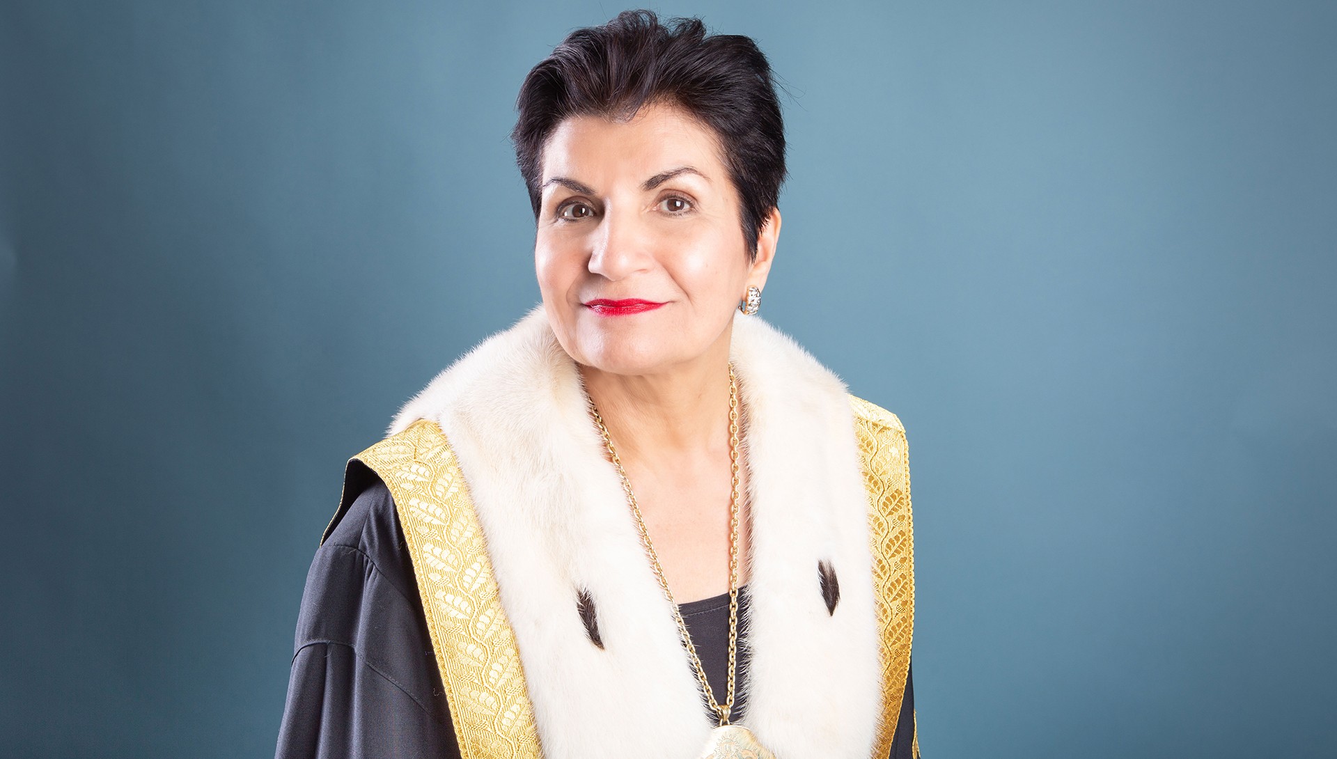  A person with short dark hair wearing a dark robe with gold embroidery and white fur trim, a gold necklace, and red lipstick, posing against a teal background.