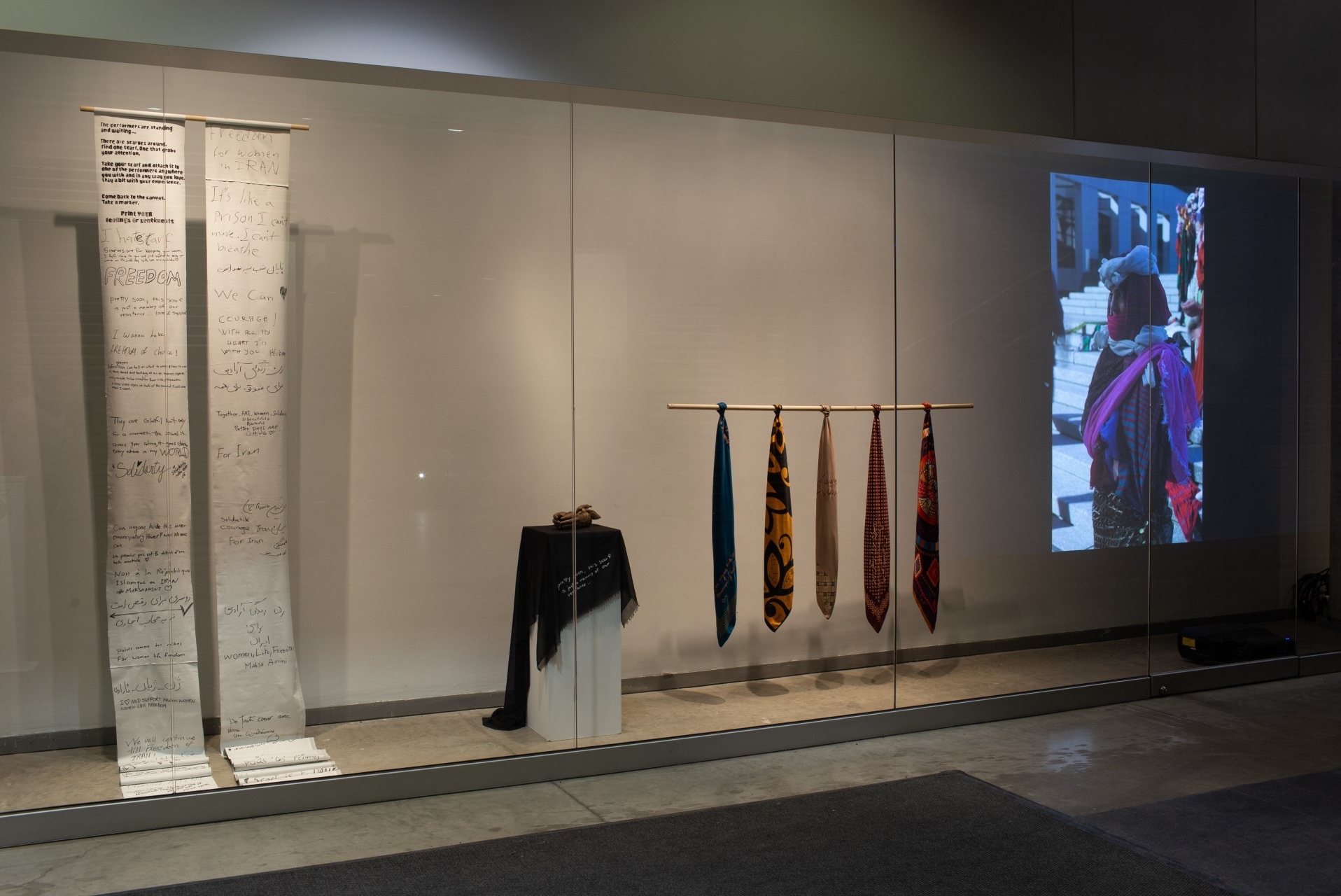 Installation view of This is not a scarf featuring a projection for a video performance, bronze casted hands, scrolls with written testimonies from the performance participants, and silkscreened scarves. 