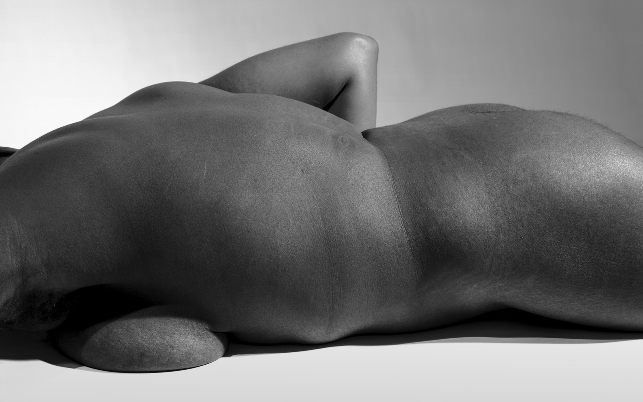 Black and white portrait of a trans identifying person lying naked on their stomach. Their body is presented like a landscape. Their face is not visible. 
