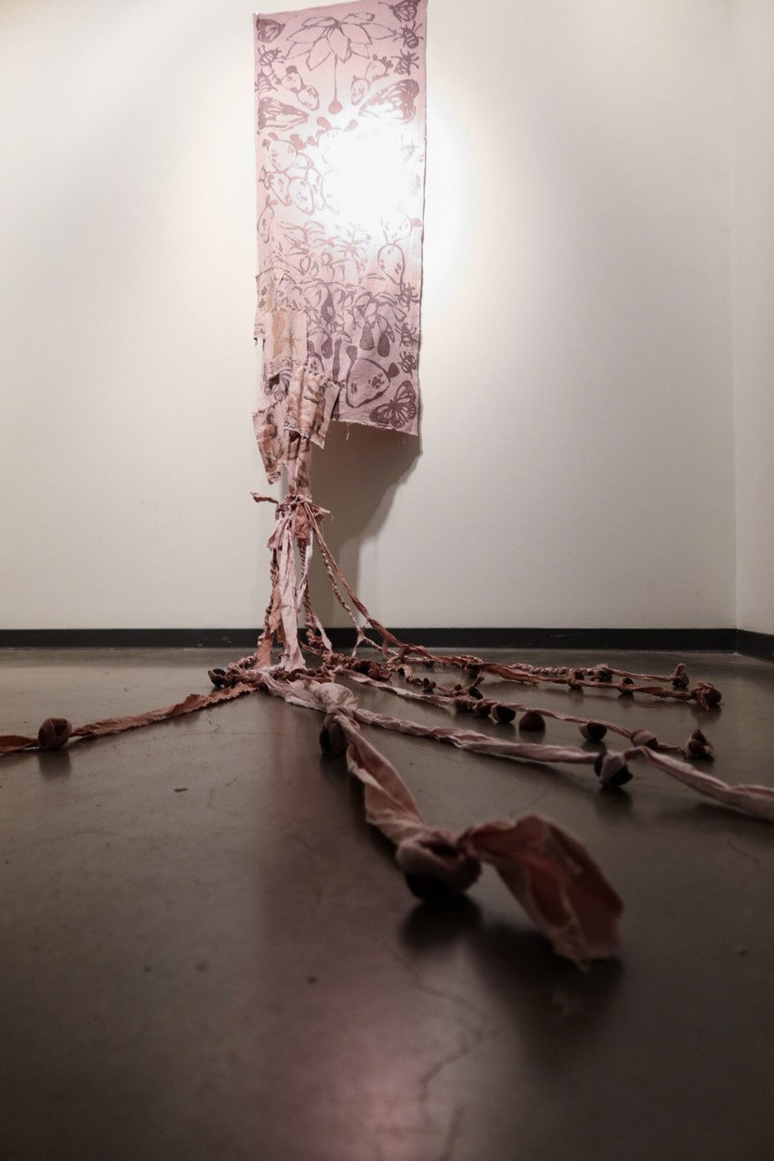 cochinilla/aguacate is a textile piece shown here hanging from the ceiling and falling to the floor with knotted fringes and beads of avocado pits. Imprints of flowers, fauna, and 18th-century illustrations of the process of making cochineal dye can be seen. The textile is of a pinkish color