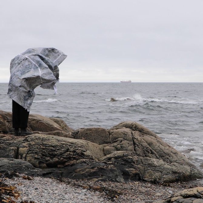 Capture vidéo,l'artiste se tien sur une surface rocheuse et est enveloppée d'une greande surface de papier au motif de roche, créature humanoïde animée, monde fantaisiste numérique