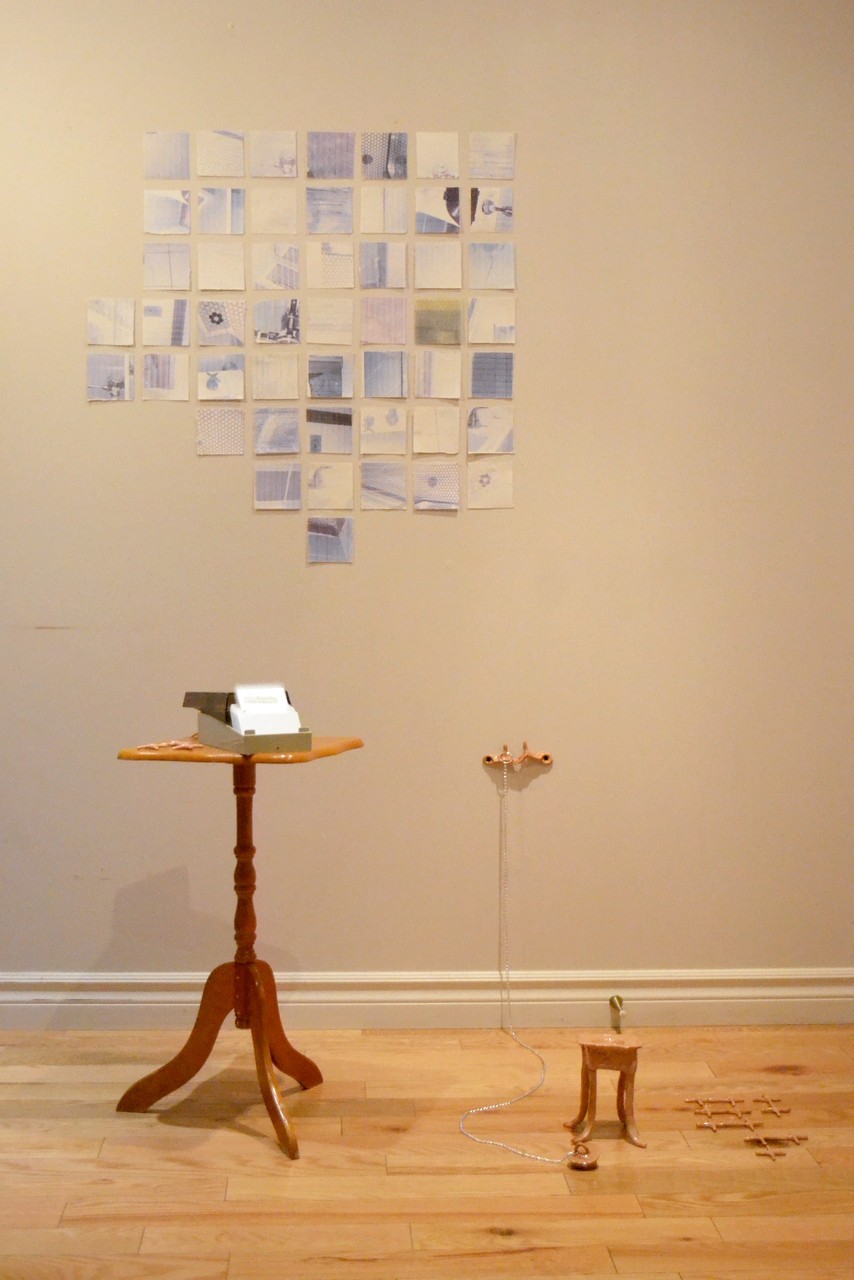 Documentation shot of the installation vestiges de nos déshabillements which includes a grid of printed images of bathroom tiles on blue-ish paper made out of gathered toilet paper sheets. In front of the wall work, there is a small table with a steel business card box with toilet paper samples and a beige ceramic cross-like shapes. Next to the table, there is a ceramic reproduction of a faucet hanging to the wall from which a chain leads to an ill-fitted bath stopper. More ceramic cross-like shapes are scattered on the floor. 