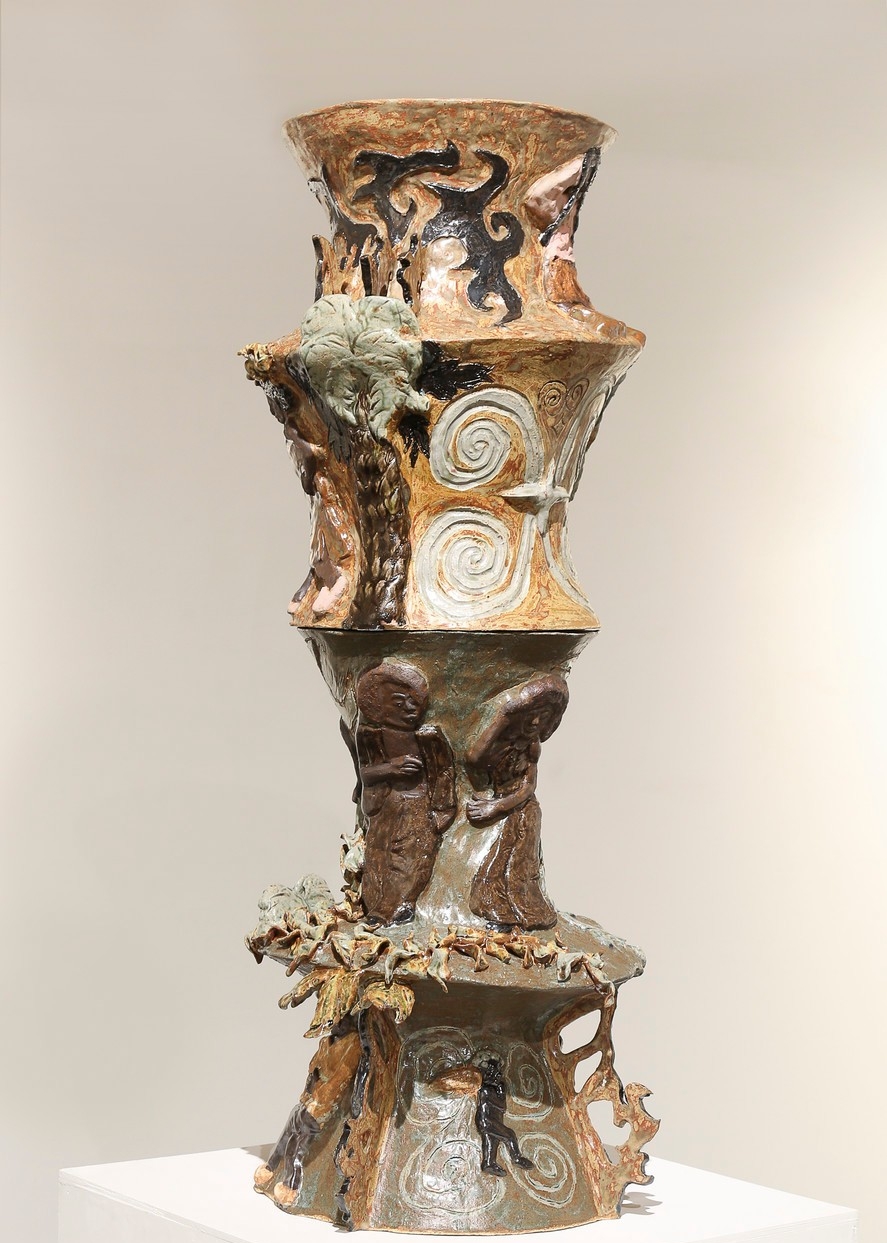 Tall ceramic sculpture with relief details and an earthy color palette standing on a plinth. The sculpture is divided in four sections vertically, looking like vases stacked on top of each other. They depict individuals who are Afro-descendant resting, dancing, and performing other forms of self-care. 