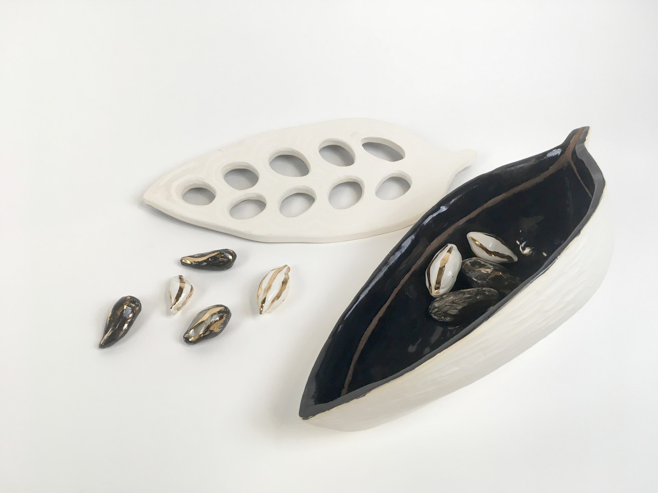 The documentation of  Seeds of Hope shows a small grouping of  small groupings of black and white glazed earthenware with gold luster displayed outside and inside the cacao bean. We can see the interior of the bean which is black with gold luster streaks and its white matte exterior. Along side the ceramic vessel there is a white matte ceramic leaf shaped piece with holes echoing the shape of the beans. 