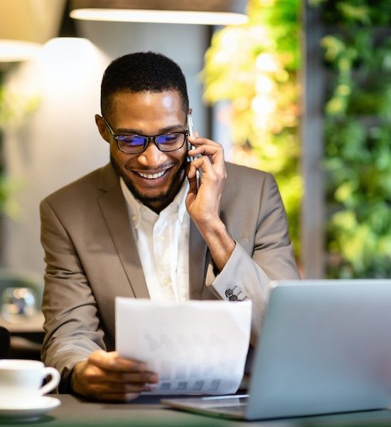A man in a suit, smiling and talking on the phone, holds a document in one hand with a laptop and a cup next to him in a modern cafe setting.