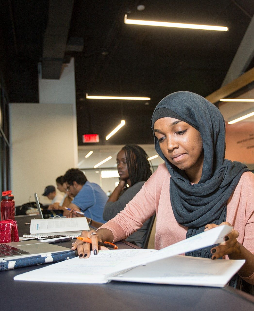 A student wearing a hijab reads a page in a notebook and other students study nearby