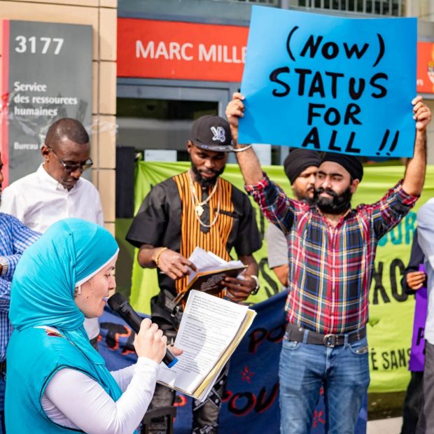 Photographie d'un groupe de manifestants, dont l'un parle dans un microphone tandis qu'un autre tient une pancarte « Un statut pour tous ».