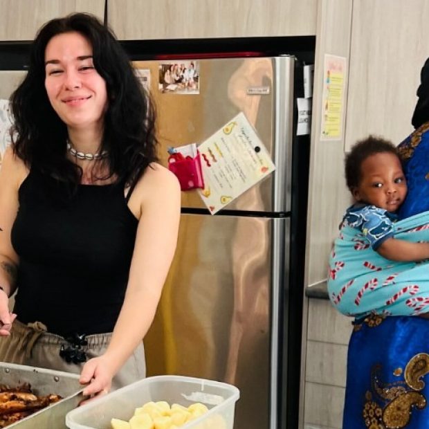 Photographie de deux femmes dans une cuisine préparant un repas copieux, tandis qu'un bébé se trouve sur le dos de l'une d'entre elles.
