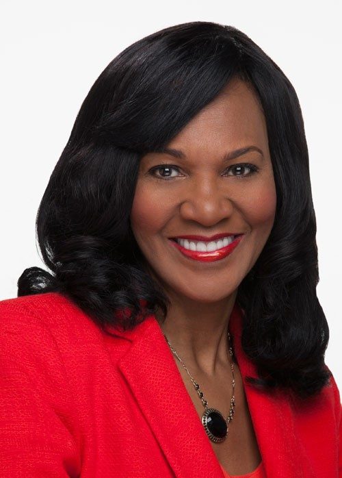 Gwen Tolbart Portrait of a smiling woman with shoulder-length black hair, wearing a red blazer, and a silver chain with black pendant