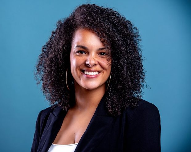 Teeanna Munro A woman with curly dark hair is smiling and wearing a dark blazer over a white tank top