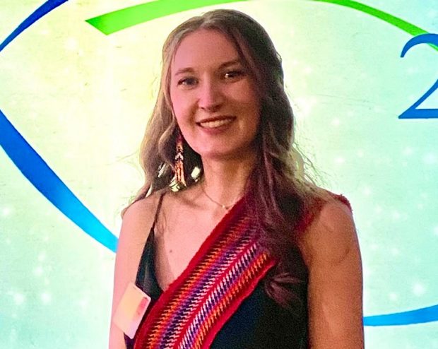 Madeline Montgomery Portrait of a smiling woman with long, blonde hair, wearing beaded earrings, a black sleeveless dress and a brightly-coloured sash