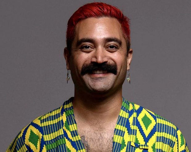 Léuli Eshrāghi A headshot of a person smiling with short dyed red hair and a moustache wears a yellow and green print shirt