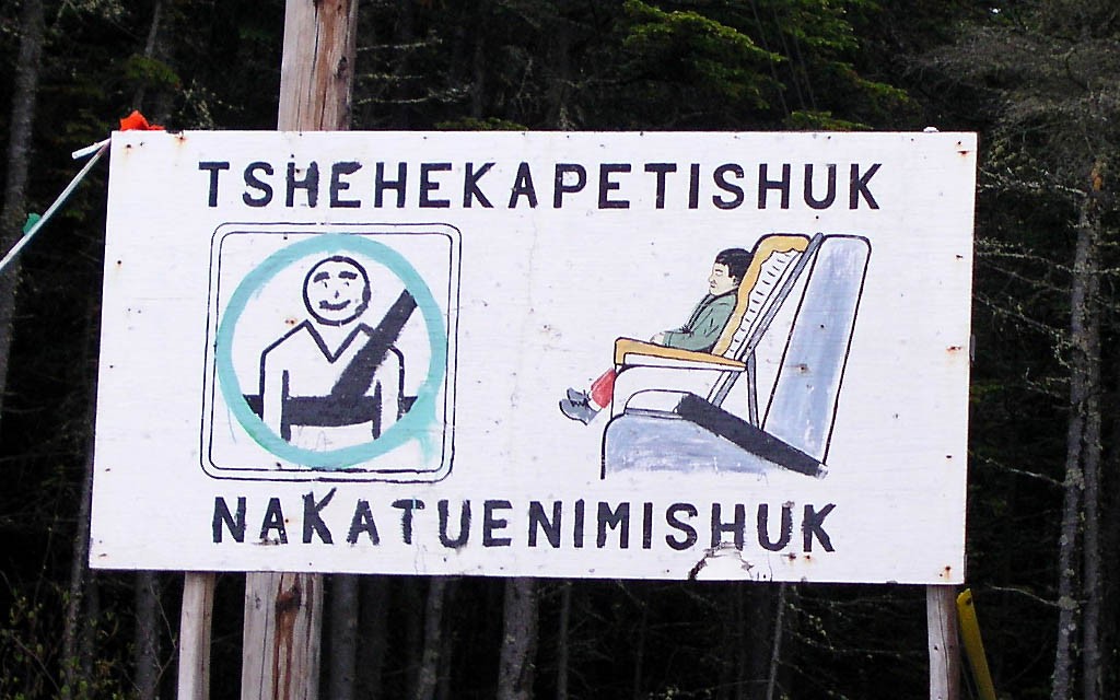 Un panneau « Port obligatoire de la ceinture de sécurité » peint à la main, avec l'inscription d'une traduction en innu-aimun « Tshehekapetishuk nakatuenimishuk » | A hand-painted “Mandatory seat belt use” sign, with the Innu-aimun translation “Tshehekapetishuk nakatuenimishuk”