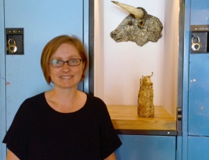 A photo of a woman with chin-length light hair and glasses standing in front of a blue wall of lockers with one locker open and a sculpture of am animal and vessel in the open locker