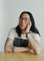 A photo of an man with straight long black hair and glasses, wearing a white t-shit with a black square, leaning his elbows on a table against a white background