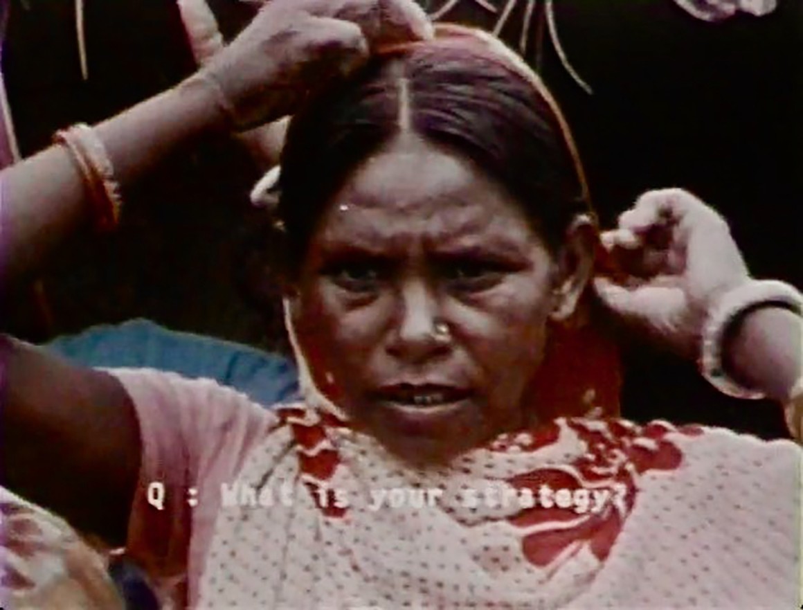 Indian woman wearing loose red-patterned garment with hands on hair and ear.