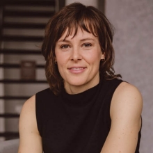 Portrait of a smiling woman in glasses, dressed in a structured floral jacket against a pink background.