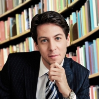 A man wearing suit and tie sits with library shelves in the background