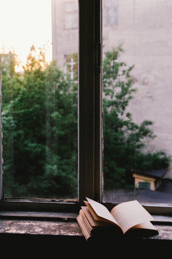 Open book in front of a window with greenery and a building visible through the window.