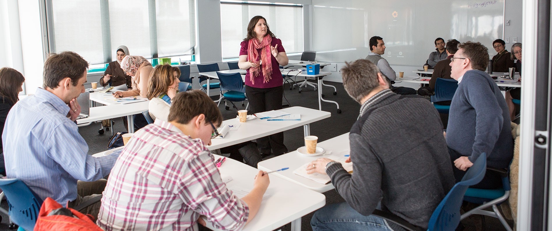 A teacher at the center of a classroom and students engaged in activities and conversations