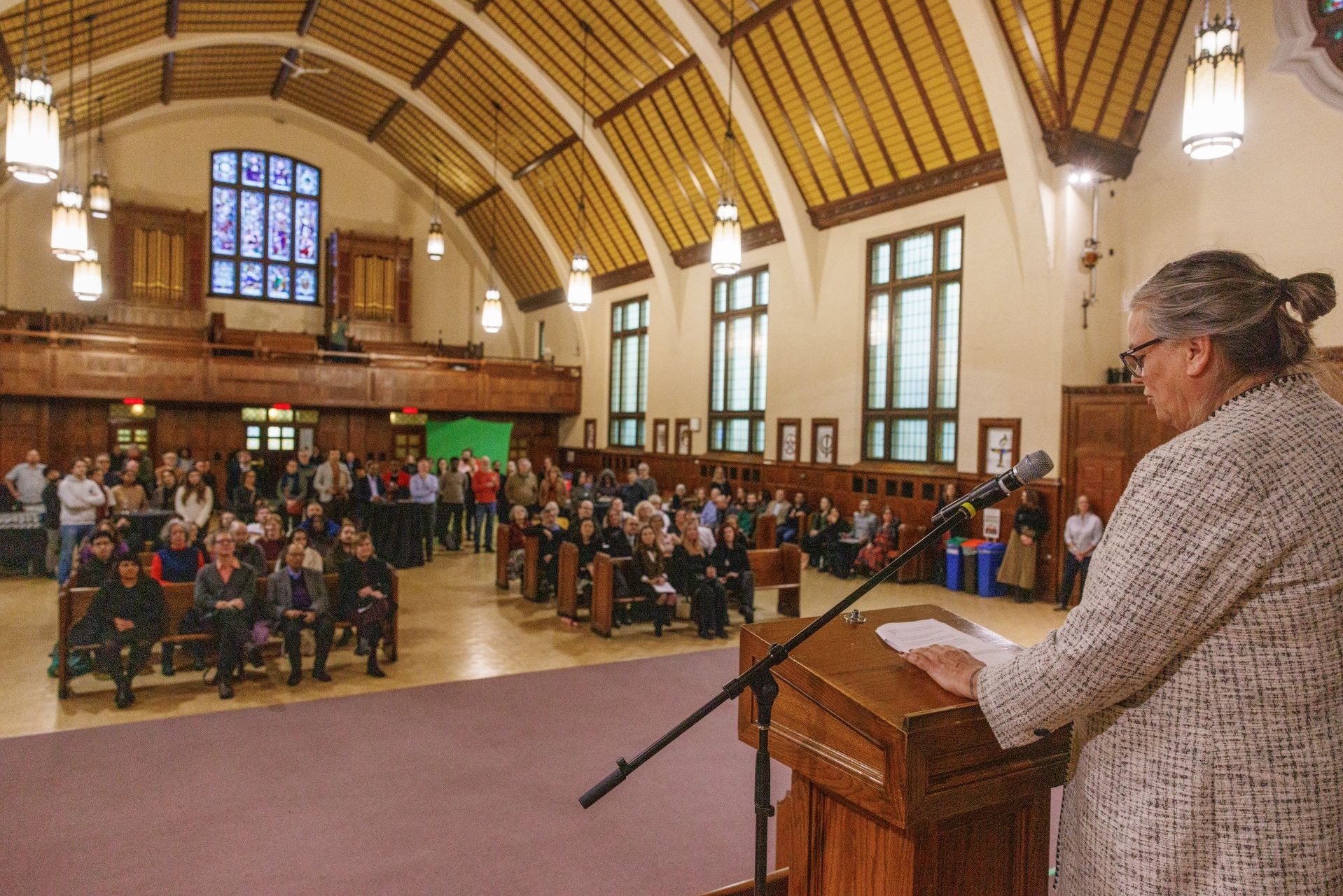 The back of a woman wearing a grey suit jacket standing at a wooden podium in front of a crowd. The crowd is sitting on chuch benches, in a chapel. 