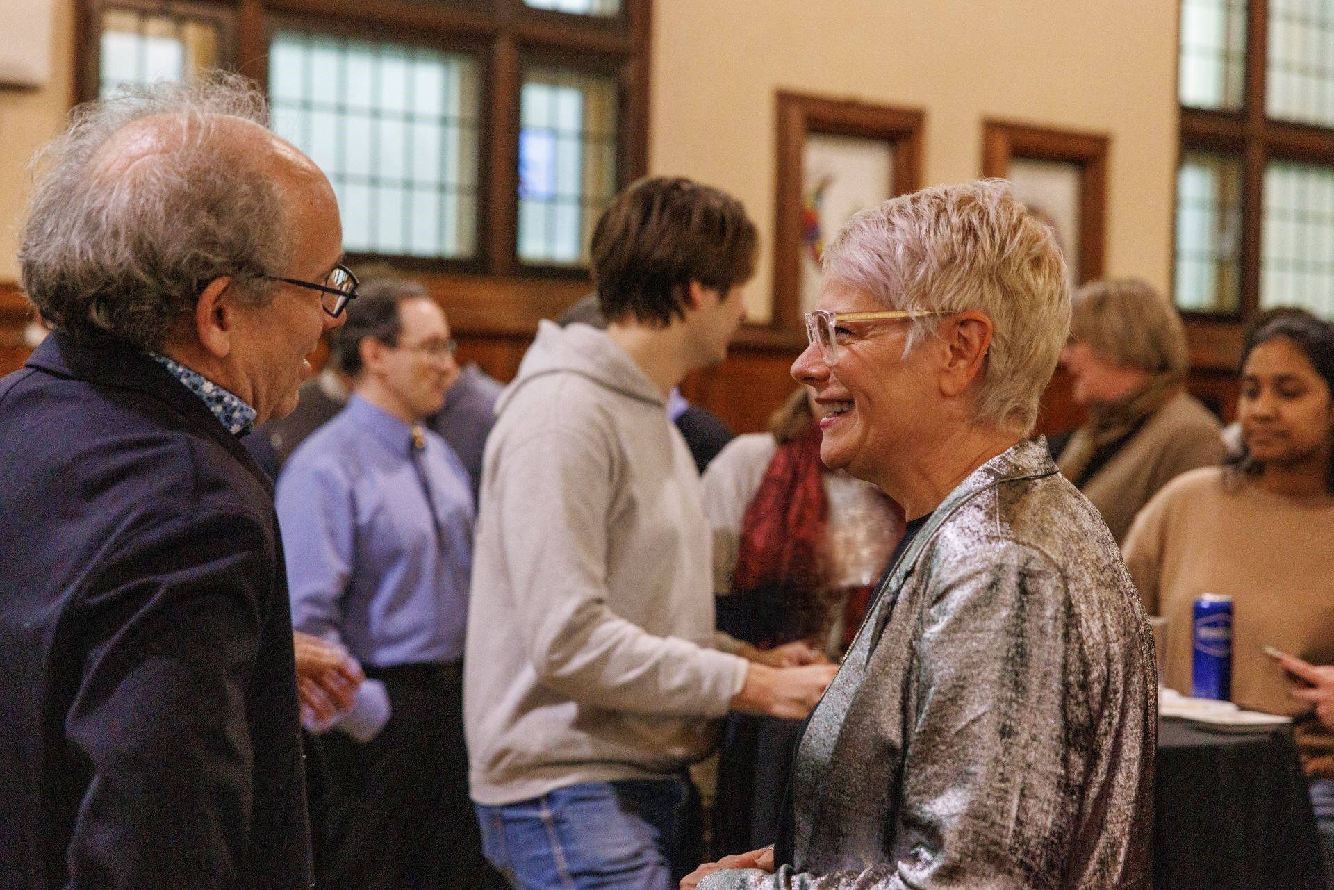 A man wearing glasses and a dark blue blaser and a woman with glasses and a silver suit jacket are smiling and having a conversation among a crowd.