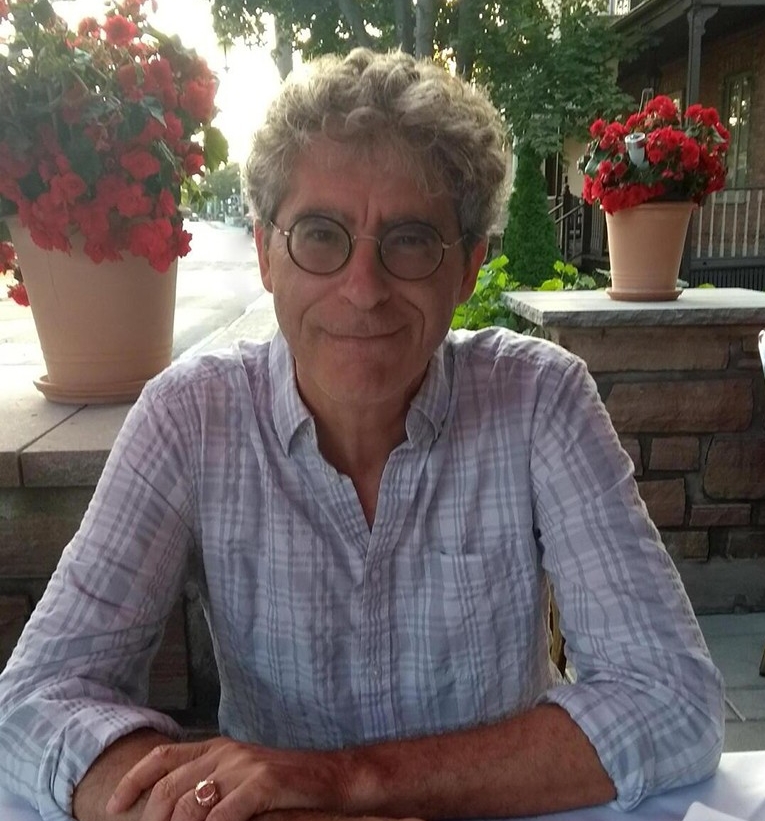 Man with curly hair and round glasses sitting outside, smiling.