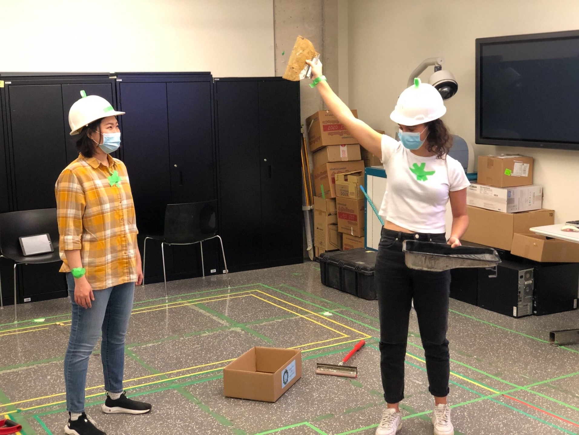 Two individuals wearing white hard hats, face masks, and casual clothes are in a room with black cabinets and boxes, engaging in an activity where one person holds a paint tray and raises a piece of material, with the floor marked with green tape.
