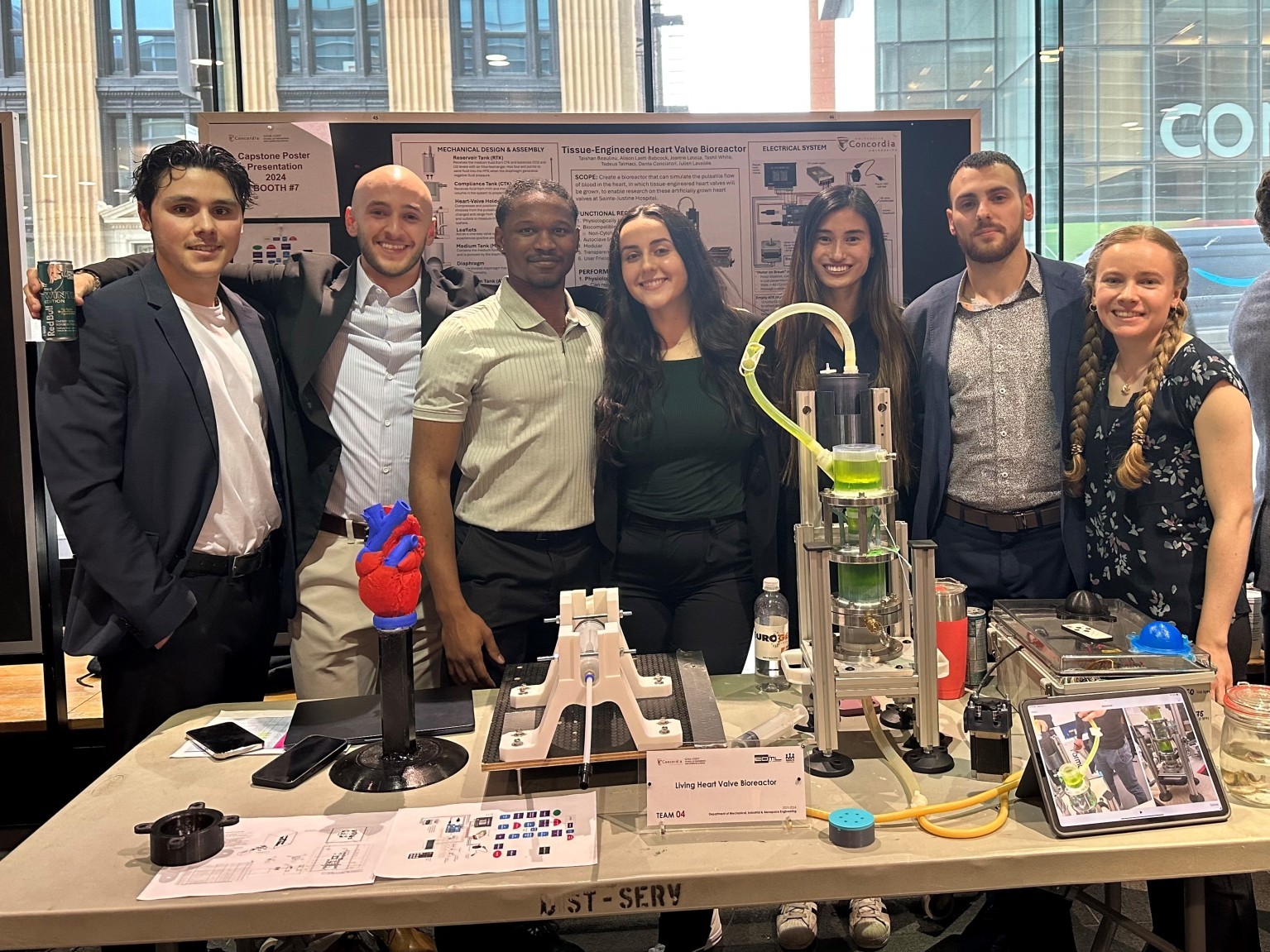 A group of seven undergraduate students stand next to one another in front of a poster and behind a table with on it a science project featuring a living hearth valve bioreactor.