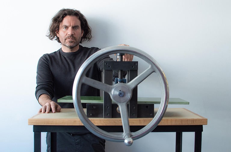 A serious-looking man with chin-length curly hair and a black top, standing behind a printmaking press.