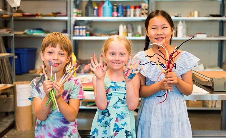 Three children smiling for the camera, holding paintbrushes and pipe cleaners
