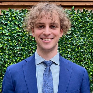 Smiling young man with curly blond hair, wearing a blue dress shirt, a blue tie and a blue blazer.