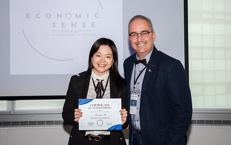 A young smiling woman holding a certificate and standing next to smiling older man in glasses and a suit.