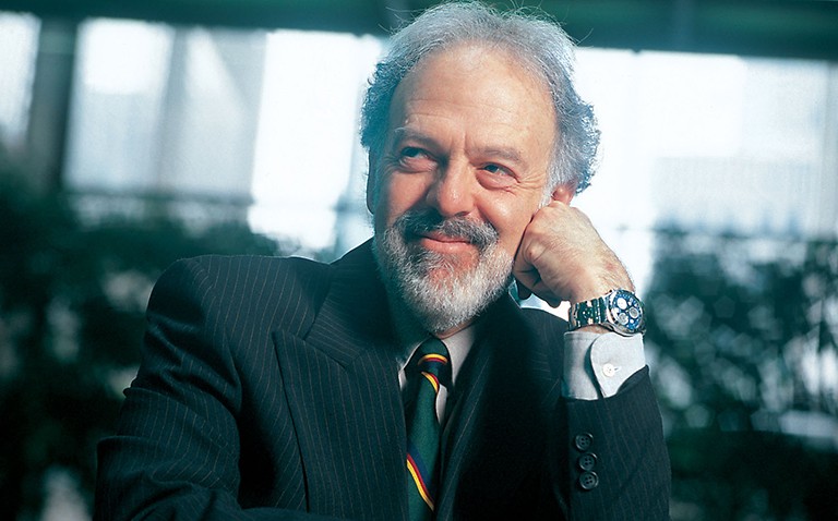 Smiling man with short, curly gray hair and a beard, wearing a black, pin-striped suit and a tie.