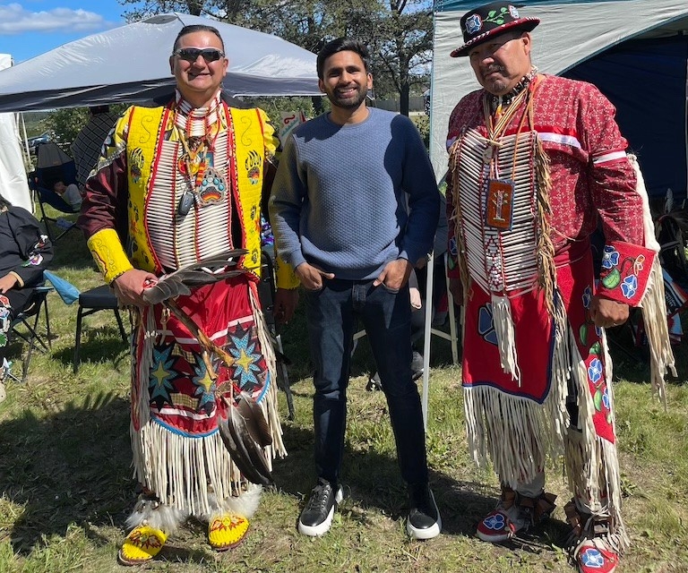 A young man stands with two Cree elders in traditional dress at a pow wow.