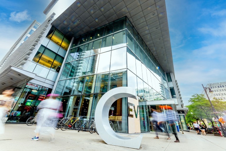 A modern glass building with a large "C" sculpture in front, surrounded by bicycles and people walking past.