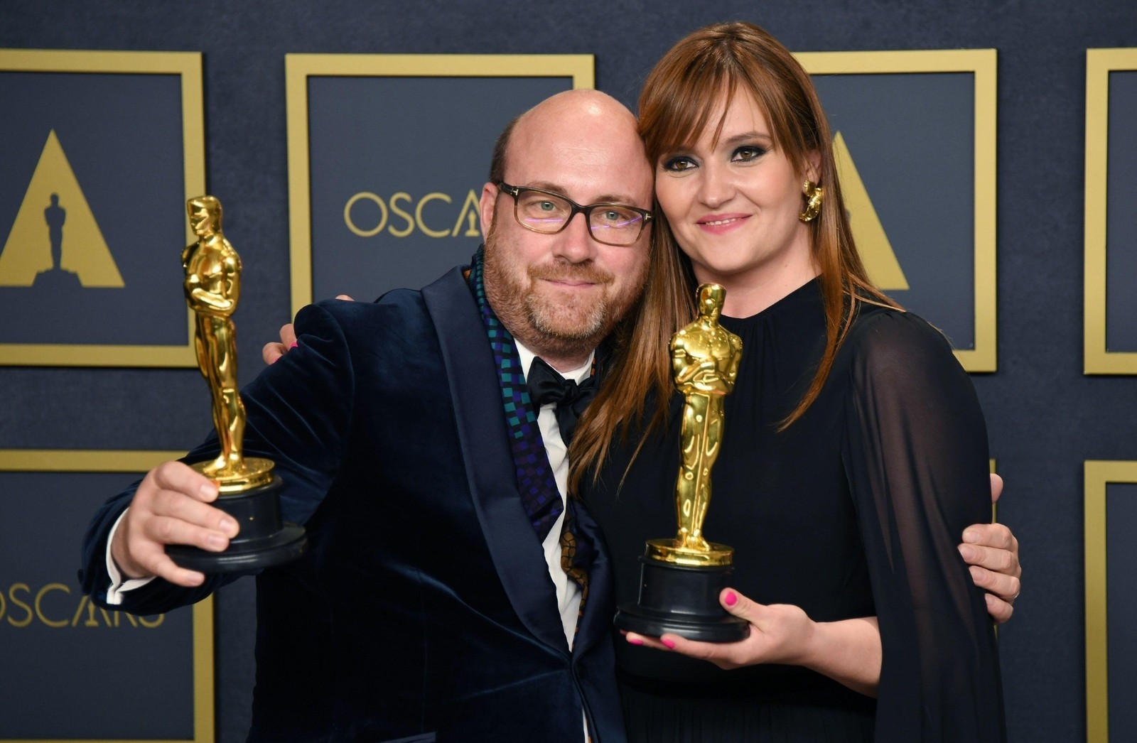 A man and a woman pose for a photo. They are dressed up and are each holding an Oscar Award in their hand.