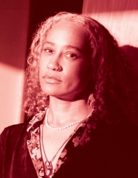 Woman with long, light, curly hair leans on a wall and looks at the camera. She wears two necklaces and a top with a floral print collar.