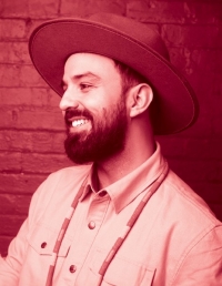 He stands before a brick wall, and wears a brimmed hat, beaded necklace, and white button-up top. He has a beard and smiles while looking away from the camera.