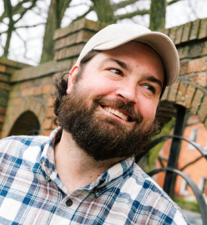 Man with a beard, plaid shirt and white baseball cap smiles in front of a brick fence.