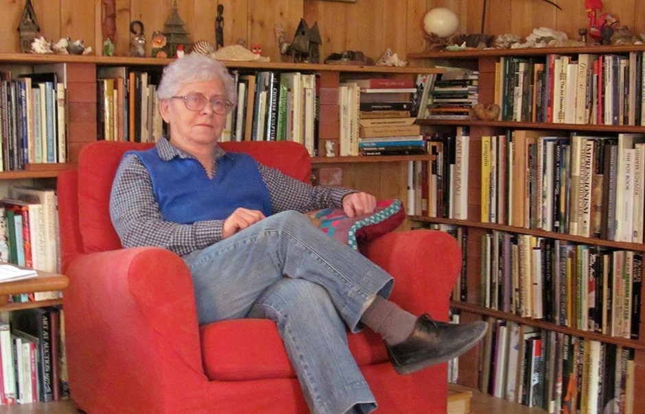 Rose has grey hair and glasses. She sits in a red chair near wooden shelves that are packed with books