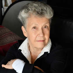 A woman with shorty grey hair wearing white collared shirt and black sweater eyes the camera.